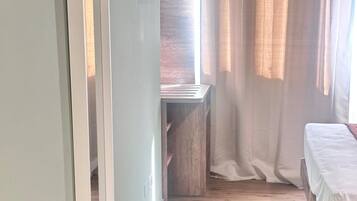 A narrow room with a wooden floor, a bed with white bedding, a green pillow, and a white pet bed with two bowls.