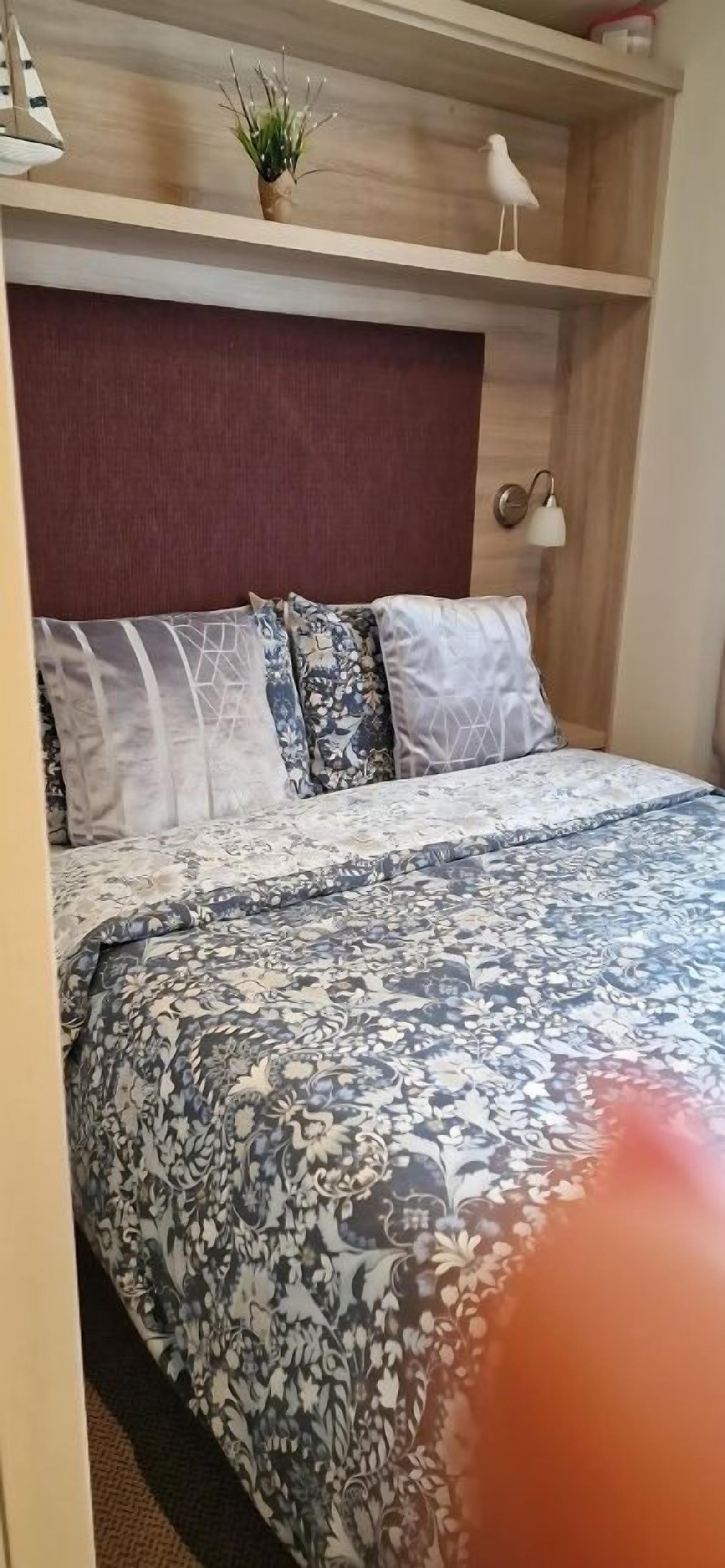 A bedroom with a bed, pillows, a headboard, a shelf with decorative items, and a wall-mounted lamp.