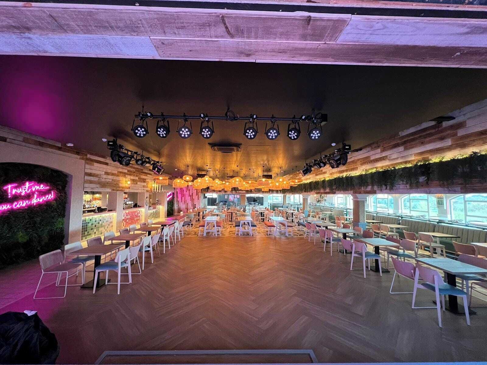 A spacious indoor dining area with multiple tables and chairs, a neon sign on the left, and a wooden ceiling with hanging lights.