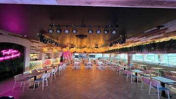 A spacious indoor dining area with multiple tables and chairs, a neon sign on the left, and a wooden ceiling with hanging lights.