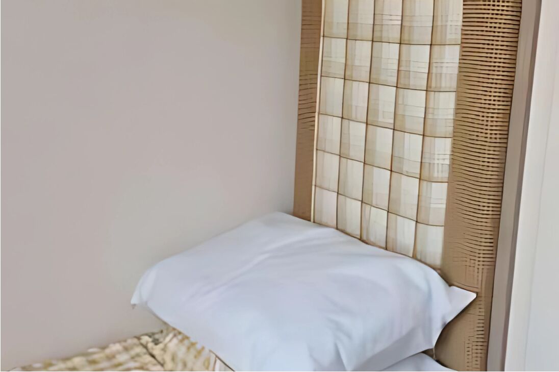 Esquina de una habitación con un almohadón blanco sobre una cama, un cabecero tejido y una pared lisa.