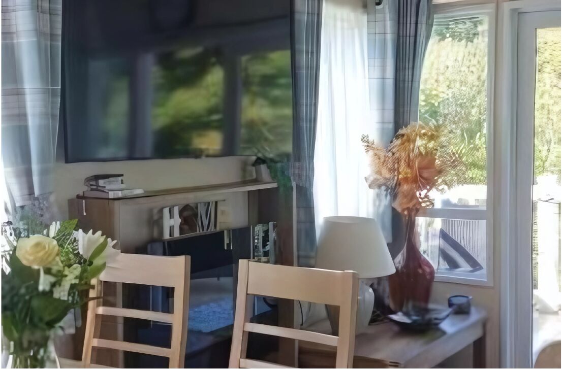 A living room with a TV mounted on the wall, a wooden table with chairs, a vase with dried flowers, and a window with curtains.