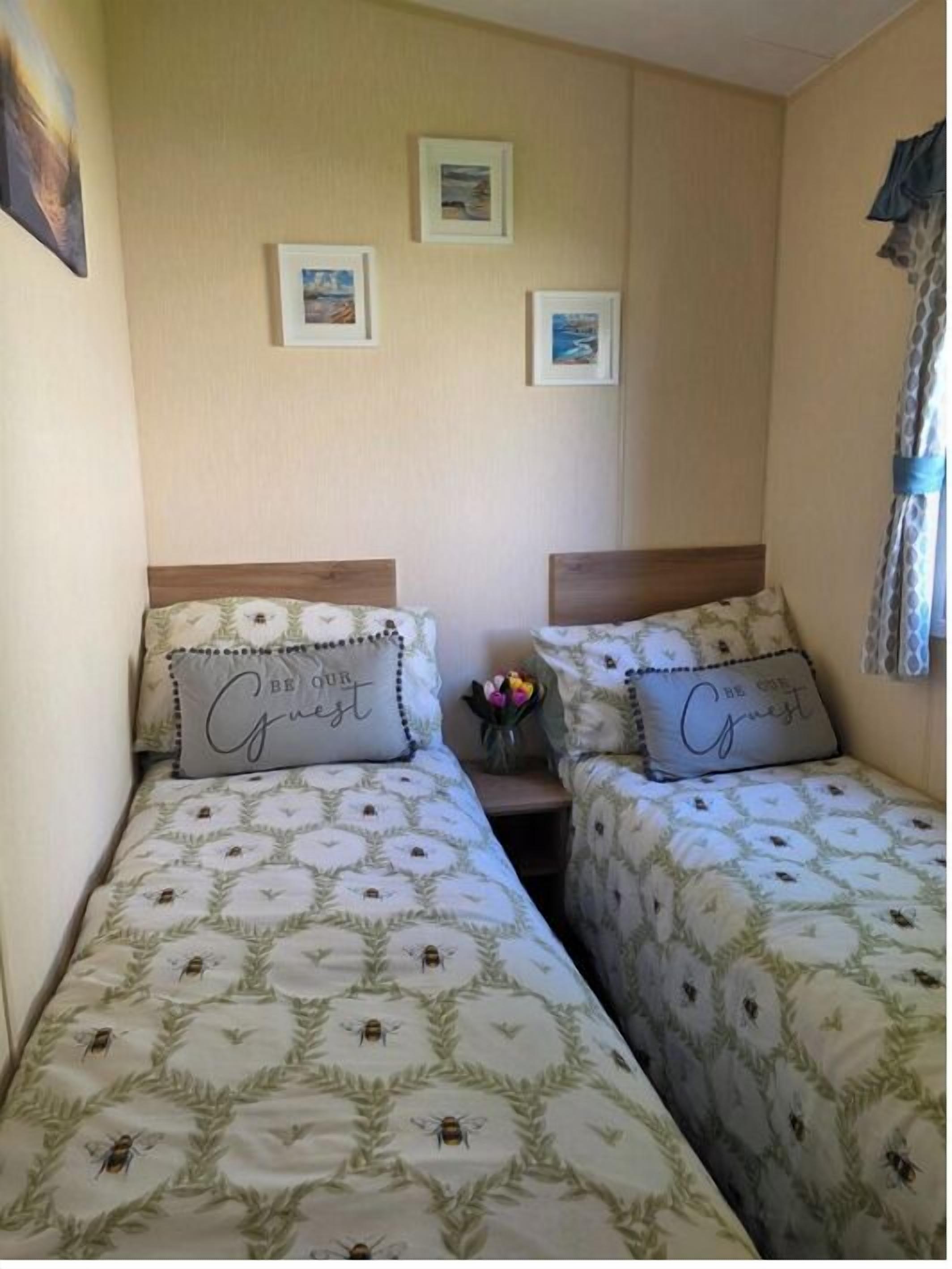 Een slaapkamer met twee bedden, elk voorzien van een bloemenprint beddeken en passend kussen. Drie ingelijste foto’s hangen aan de muur en op het nachtkastje staat een vaas met bloemen.