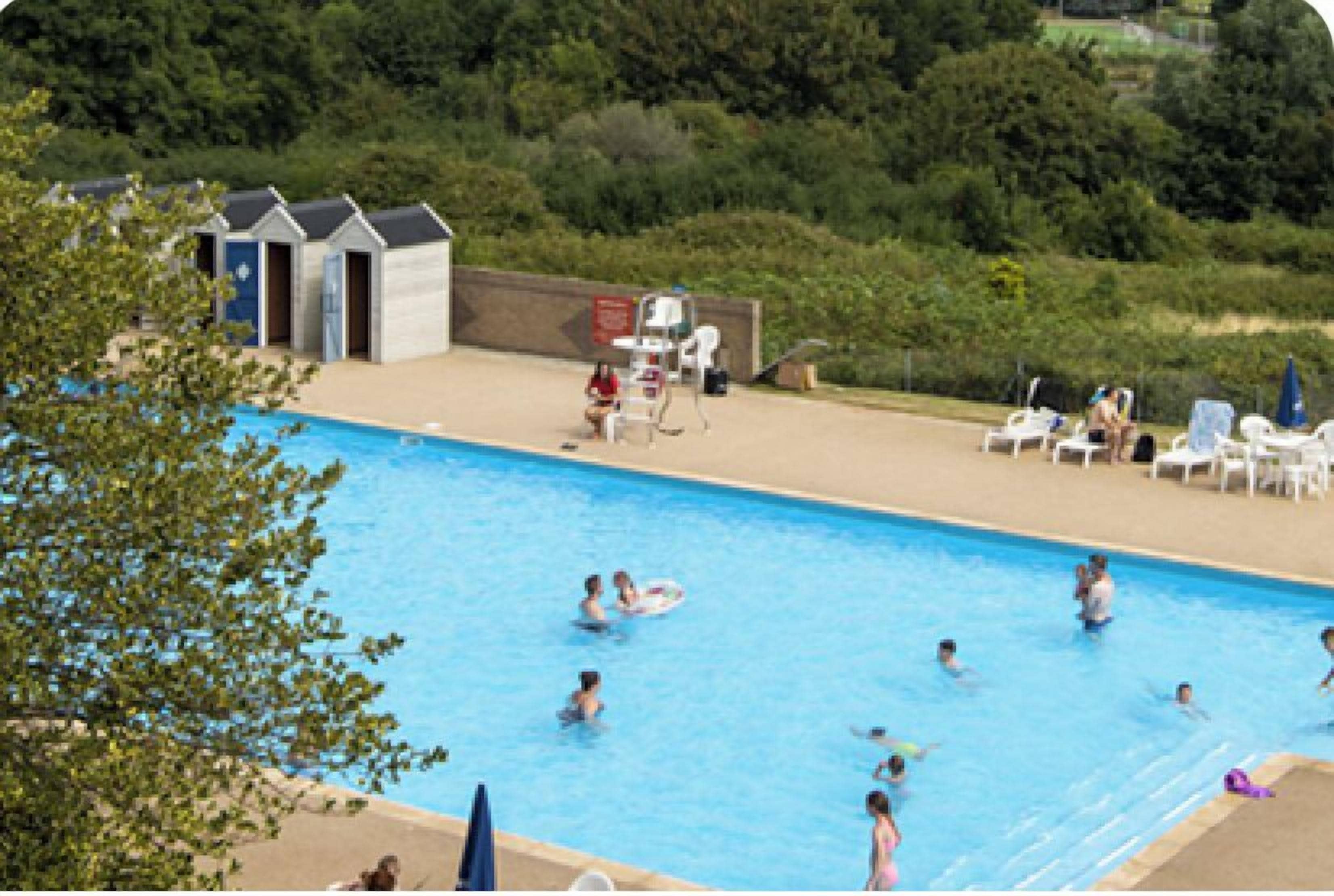 Une piscine avec des personnes dans l’eau, des chaises longues et un petit bâtiment en arrière-plan.