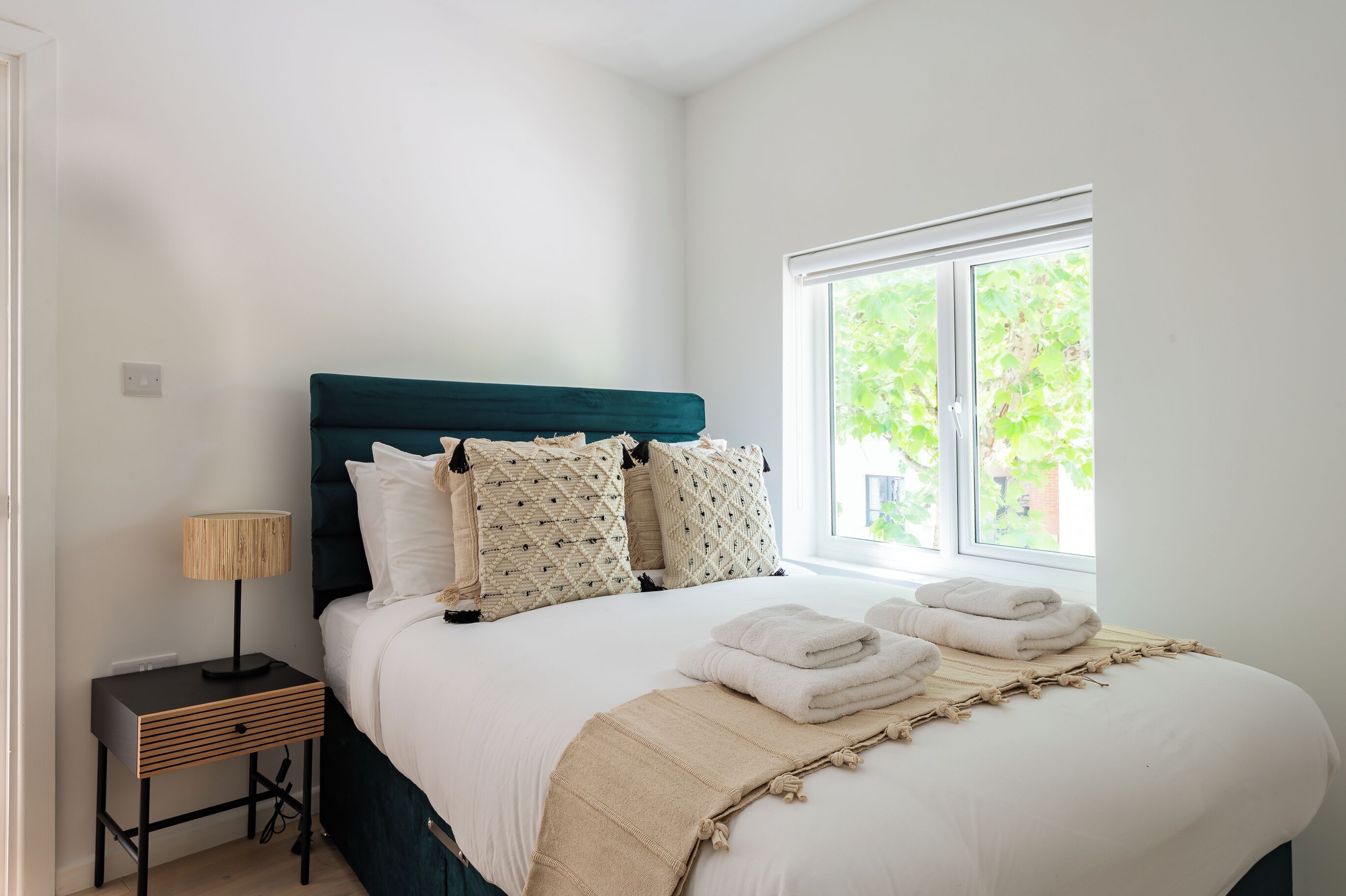 Cama bem arrumada com cabeceira verde-água, roupa de cama branca e manta bege. Há um abajur em um criado-mudo de madeira.