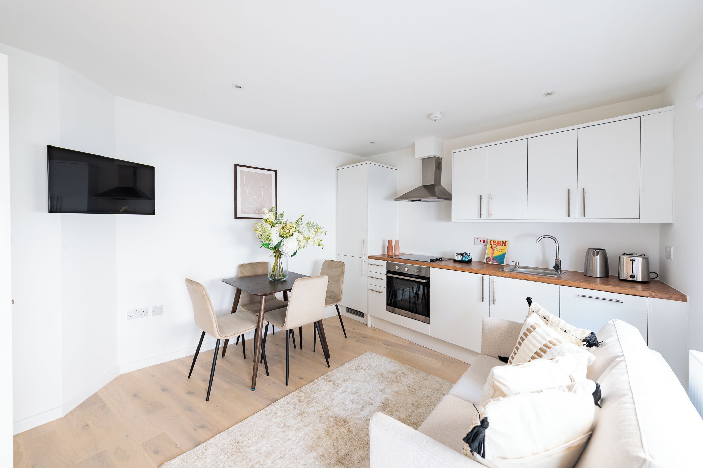 A modern kitchen with white cabinets, a wooden countertop, and a built-in oven. There is a dining area with a table and chairs, and a white sofa in the living room.