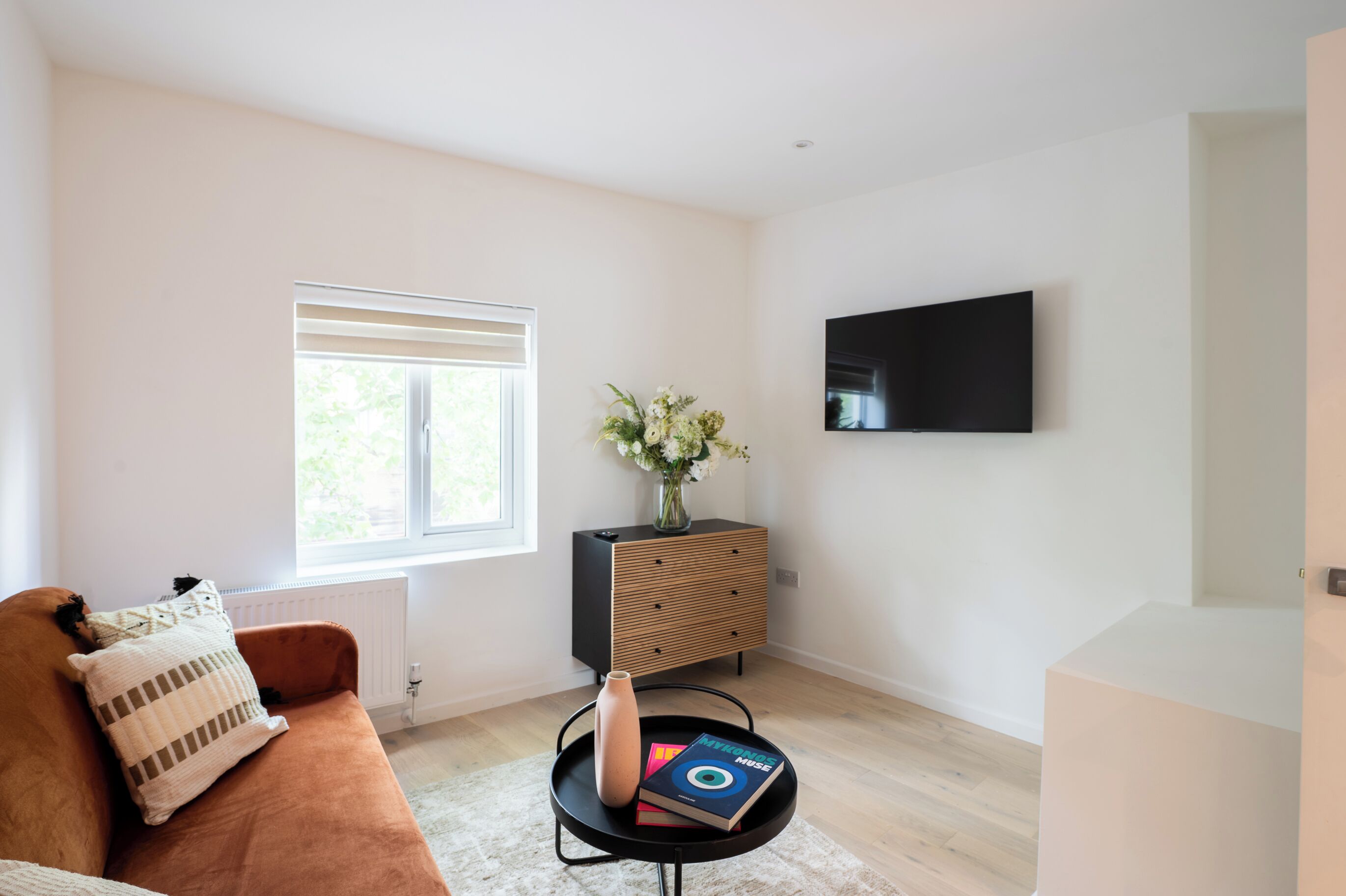 A modern living room with a brown sofa, a black coffee table with a vase and books, a wooden cabinet, and a flat-screen TV mounted on the wall.