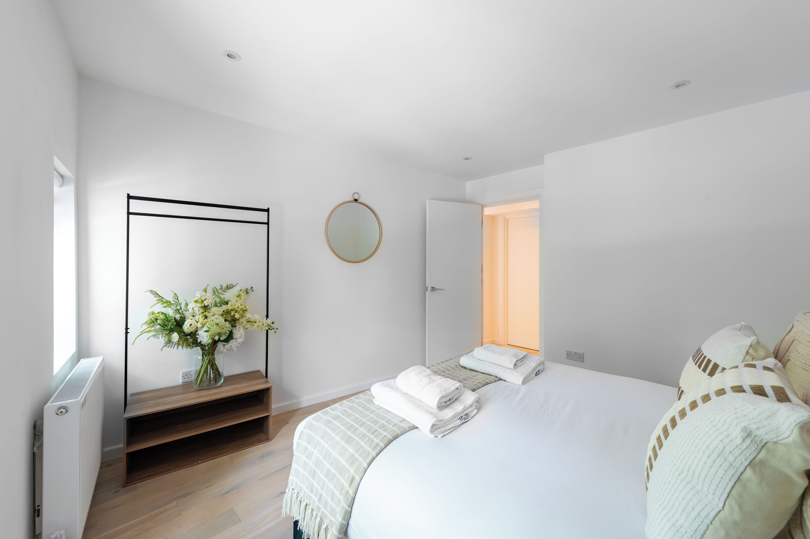 A modern bedroom with a large bed, a bedside table with a vase of flowers, a round mirror, and a door leading to another room.