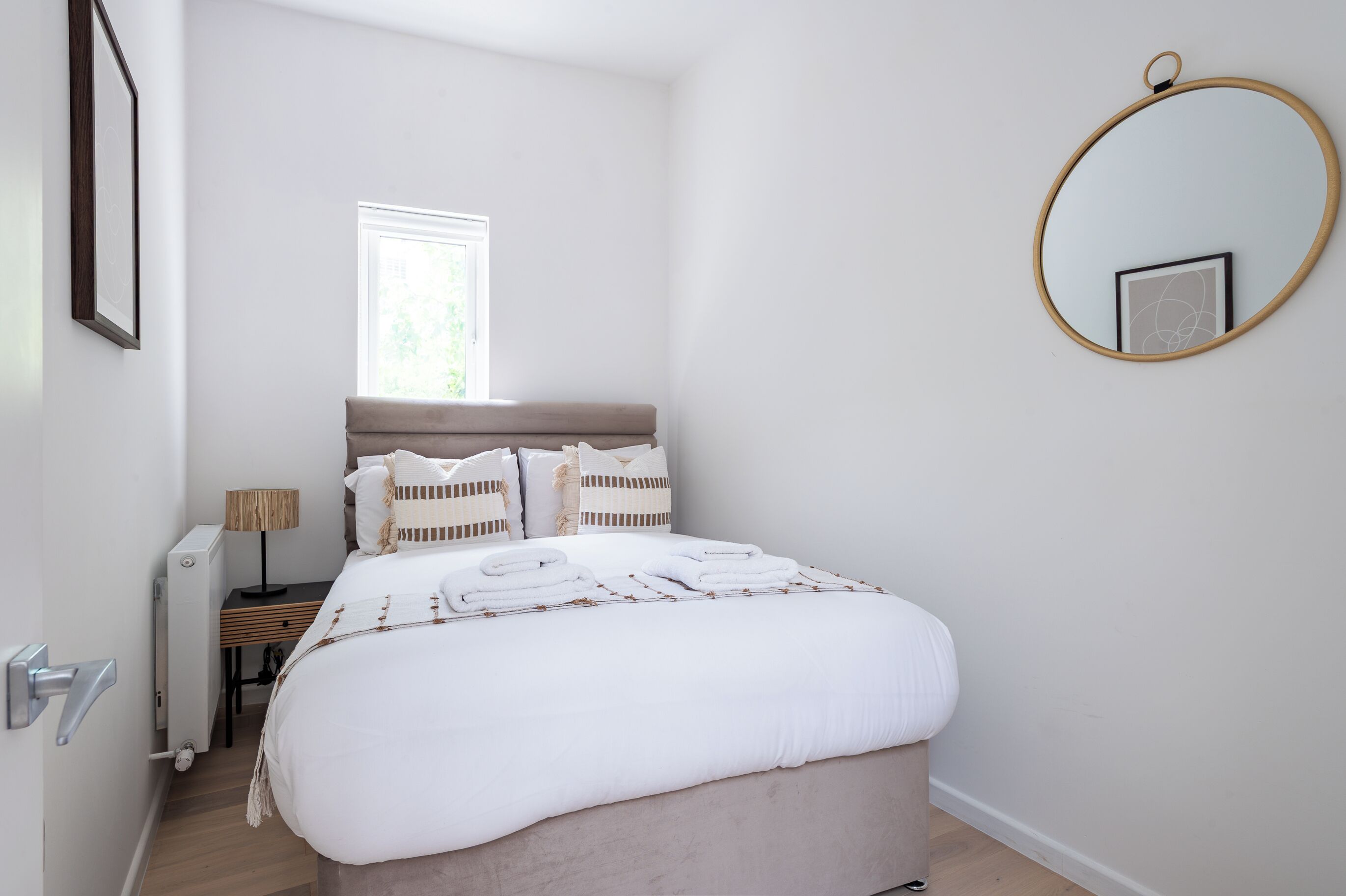 A bedroom with a bed, a bedside table with a lamp, a window, and a round mirror on the wall.