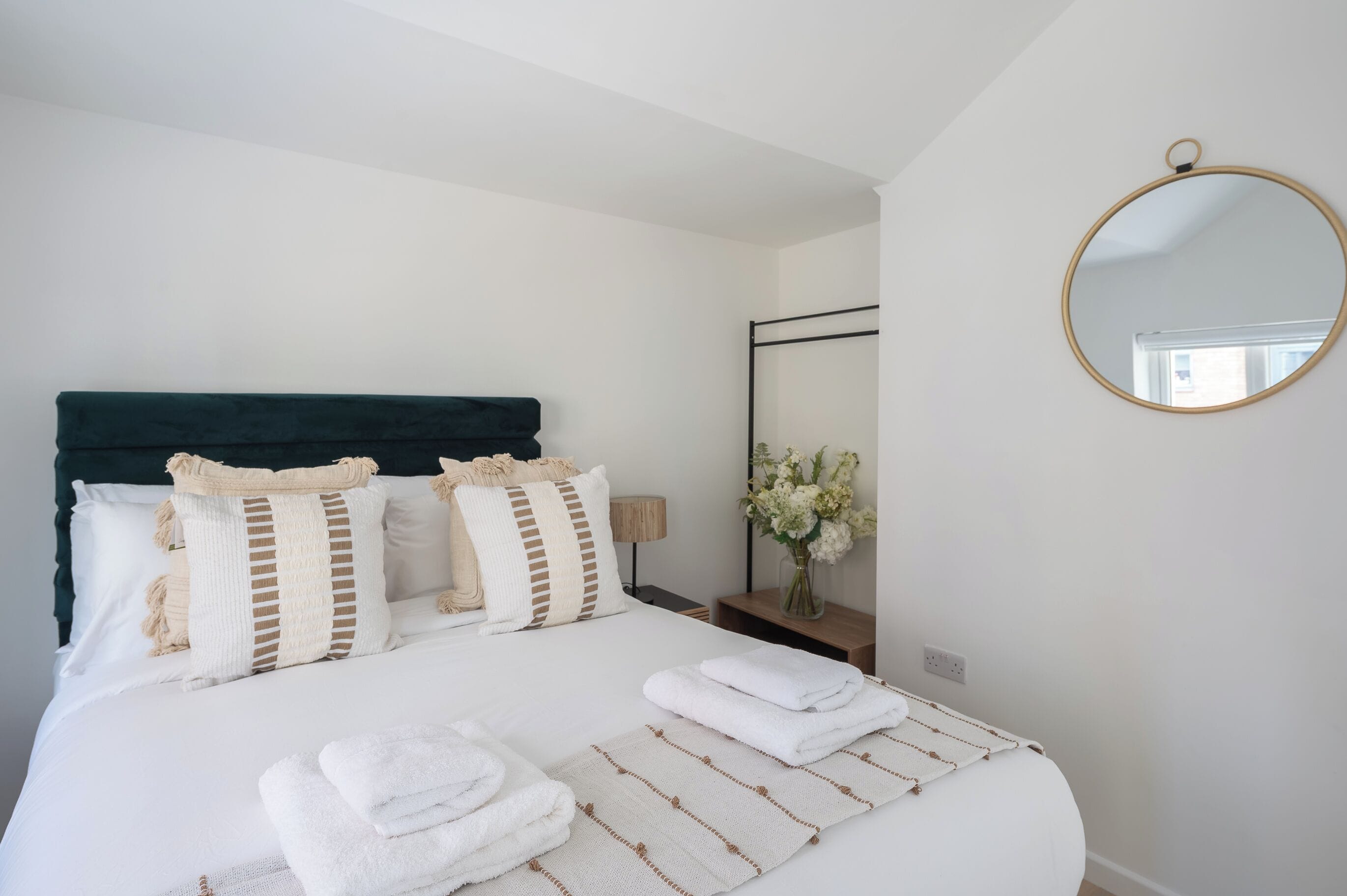 Una cama bien hecha con sábanas blancas y almohadas a rayas, una mesita de noche con una lámpara y un florero, y un espejo redondo en la pared.