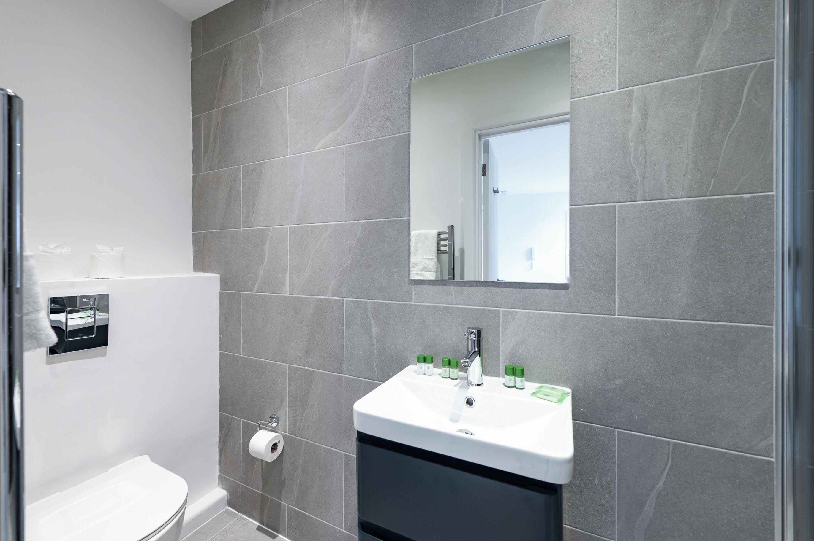 A modern bathroom with grey tiles, a white sink with green soap dispensers, a toilet, and a mirror above the sink.