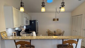 A kitchen with granite countertops, white cabinets, a microwave, stove, and a refrigerator. There are two bar stools with patterned cushions and a dining table with chairs. - Modern charm meets vibrant living with outdoor oasis and golf cart fun! (The Villages)