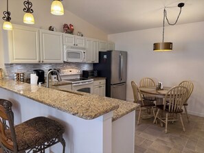 A kitchen with white cabinets, a granite countertop, a stainless steel refrigerator, a microwave, a stove, and a dining area with wooden chairs and a table.