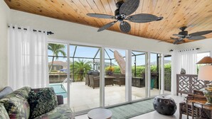 Un vaste espace de vie agrémenté d’un ventilateur de plafond, d’un canapé avec des coussins à motifs floraux, d’une table à manger avec des chaises, et offrant une vue sur une terrasse extérieure aménagée avec des meubles et des plantes, accessible par des portes vitrées.