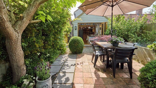 A patio with a table and chairs, surrounded by lush greenery and a tree. - Host & Stay | Romney Cottage (Corpusty)