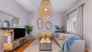 A modern living room with a television, wooden coffee table, and a sofa with a throw blanket. There are two mirrors on the wall, a hanging wicker lamp, and a window with curtains. - Green Oaks Apartment in Tamboerskloof (Cape Town)