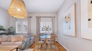 A modern living room with a sofa, coffee table, and dining table with chairs. There are windows with curtains, a hanging light fixture, and framed artwork on the wall. - Green Oaks Apartment in Tamboerskloof (Cape Town)