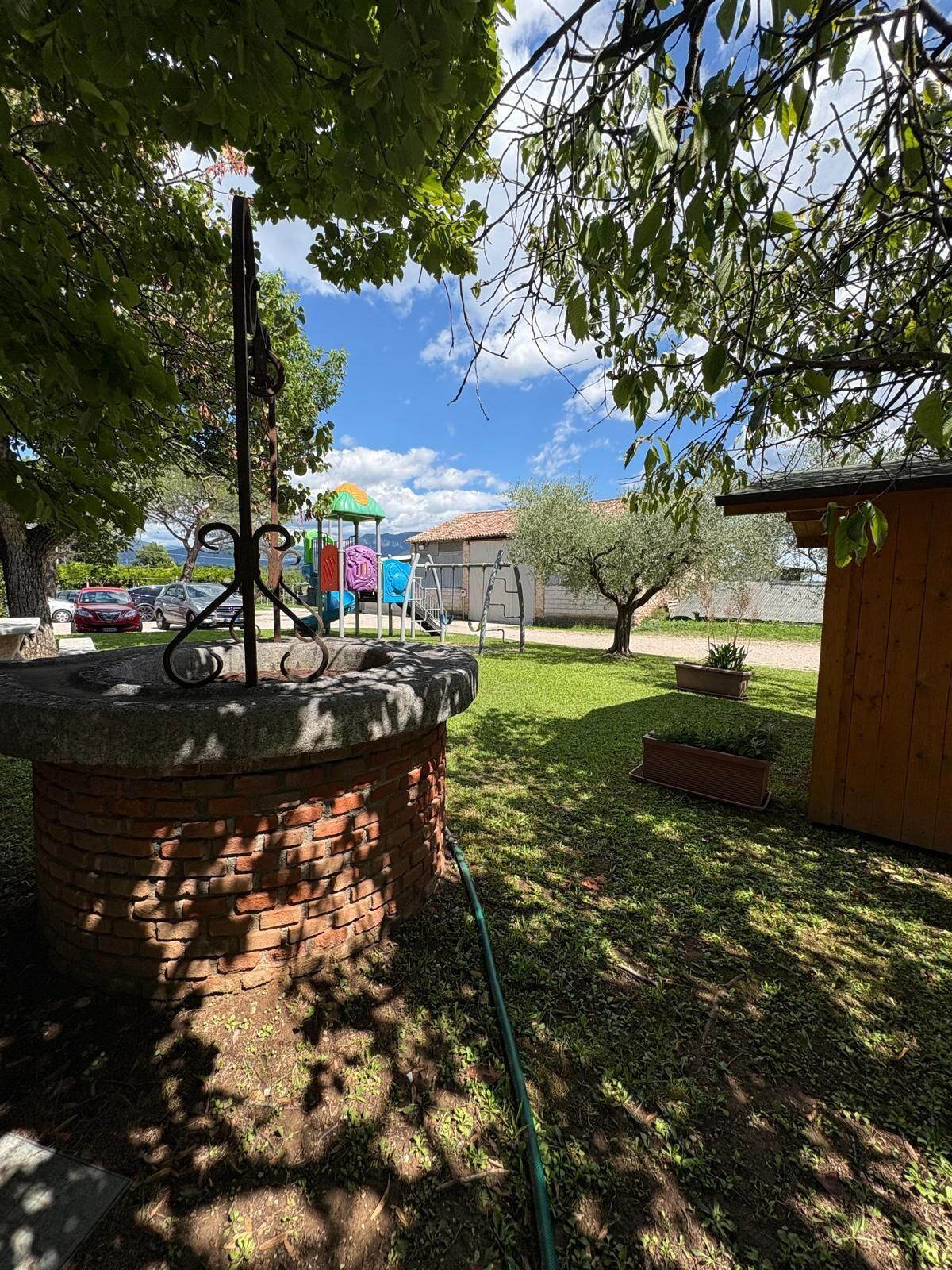 A brick structure with a metal pole, a playground with colorful equipment, and a wooden shed in a grassy area with trees.