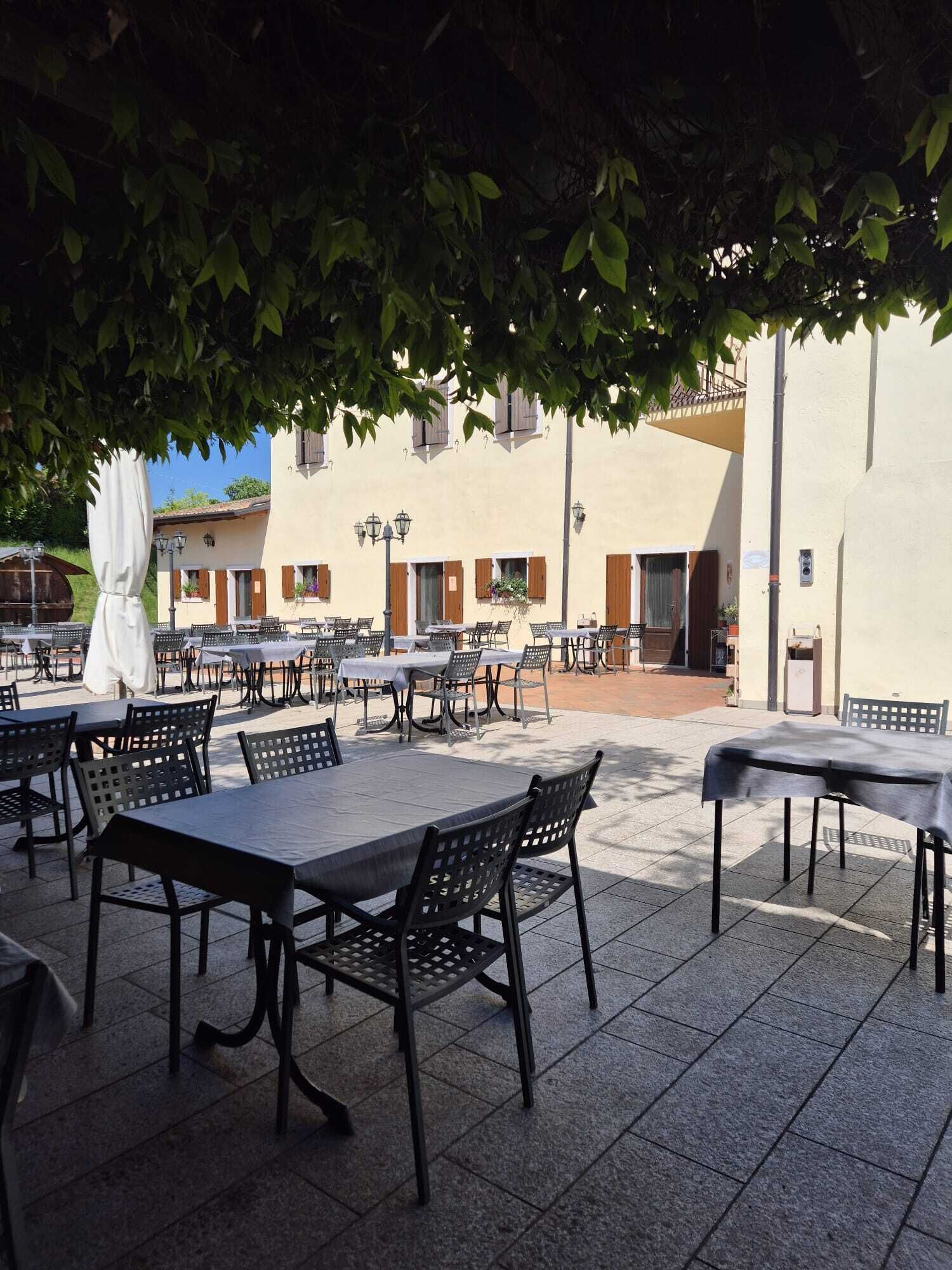 Eine Terrasse im Freien mit Tischen und Stühlen, ein weißes Gebäude mit Fenstern und ein Baum, der Schatten spendet.