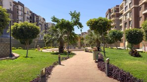 A pathway leading through a landscaped area with trees and shrubs, flanked by multi-story residential buildings.