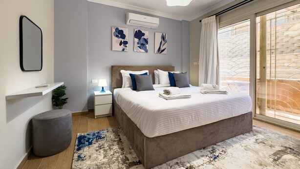 A modern bedroom with a large bed, bedside tables, a nightstand, a wall-mounted air conditioner, and a view of the outside balcony.