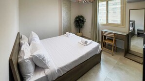 A neatly made bed with pillows and towels, a small desk with a chair, a potted plant, and a mirror. - Xuru Stays I Elegant & Modern 3 BDR Apt. @ Heliopolis (Cairo)