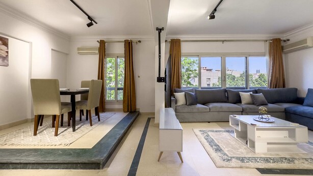 A modern living room with a grey sofa, a white coffee table, and a dining area with a table and chairs. - Xuru Stays I The 2bdr Velvet Vibe @ Maadi (Cairo)