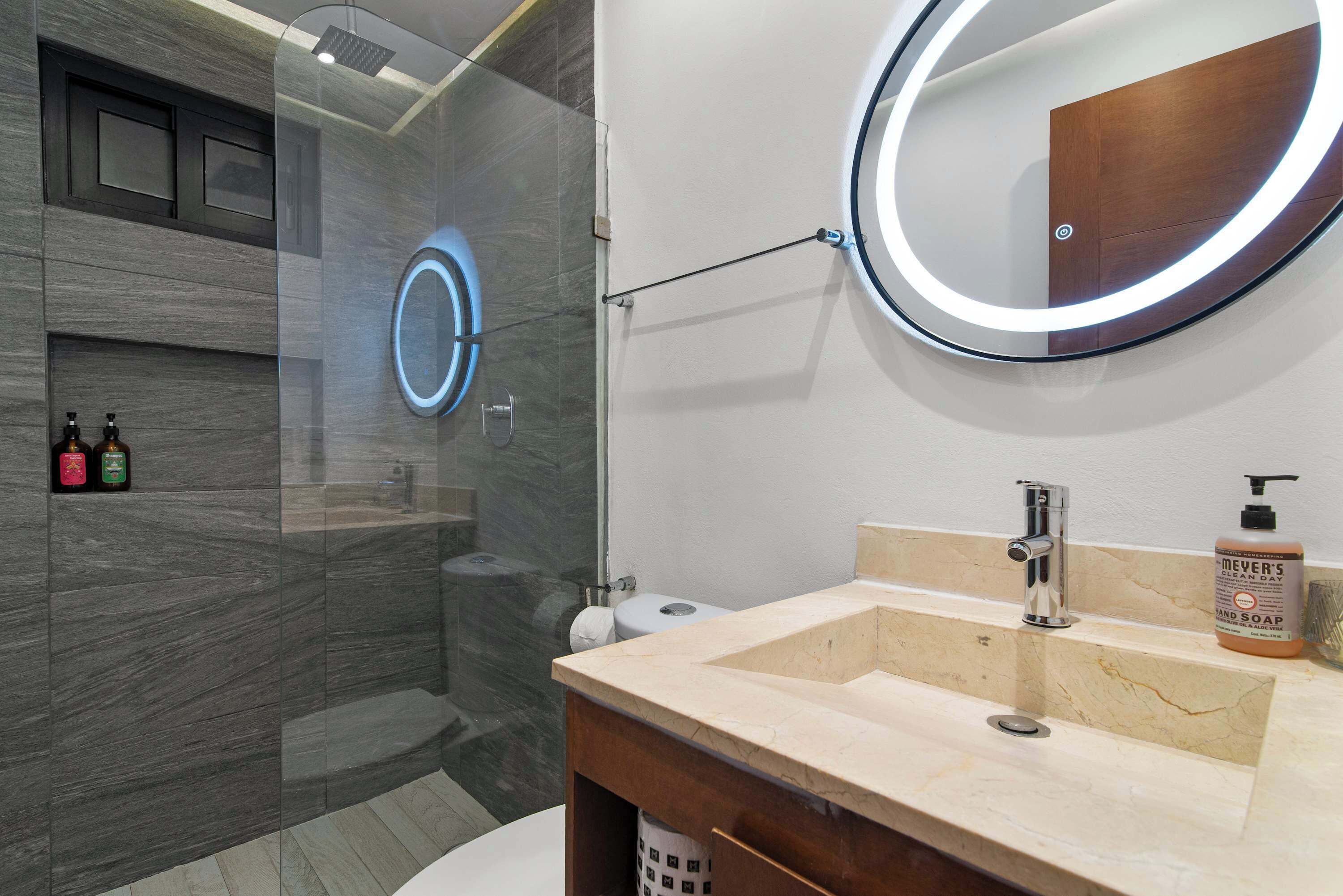 A modern bathroom with a glass-enclosed shower, a round mirror with a light, and a beige sink with a soap dispenser.