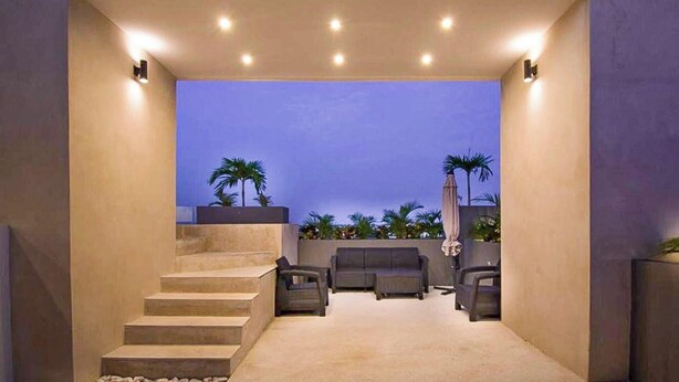 A modern outdoor living area with a staircase leading to a balcony, furnished with sofas and chairs, and surrounded by palm trees. - Stylish Studio in Zazil-Ha - Rooftop Pool (Playa del Carmen)