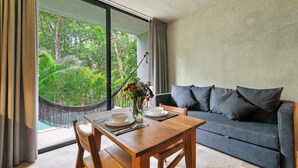 A dining area with a table set for two, a sofa, and a view of lush greenery through large windows. - Seremonia Origen - Tulum Studio with Plunge Pool (Tulum)