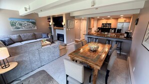 A modern living room with a grey sectional sofa, a wooden dining table with a decorative centerpiece, and a kitchen with stainless steel appliances.