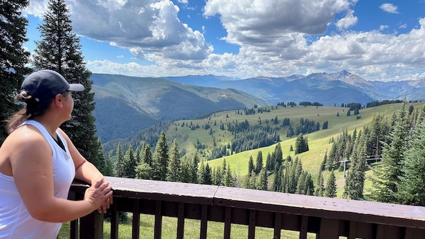 A person standing on a wooden deck overlooking a mountainous landscape with trees and a clear sky.