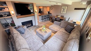 A modern living room with a large sectional sofa, a flat-screen TV mounted on the wall, a fireplace, and a kitchen area with bar stools.