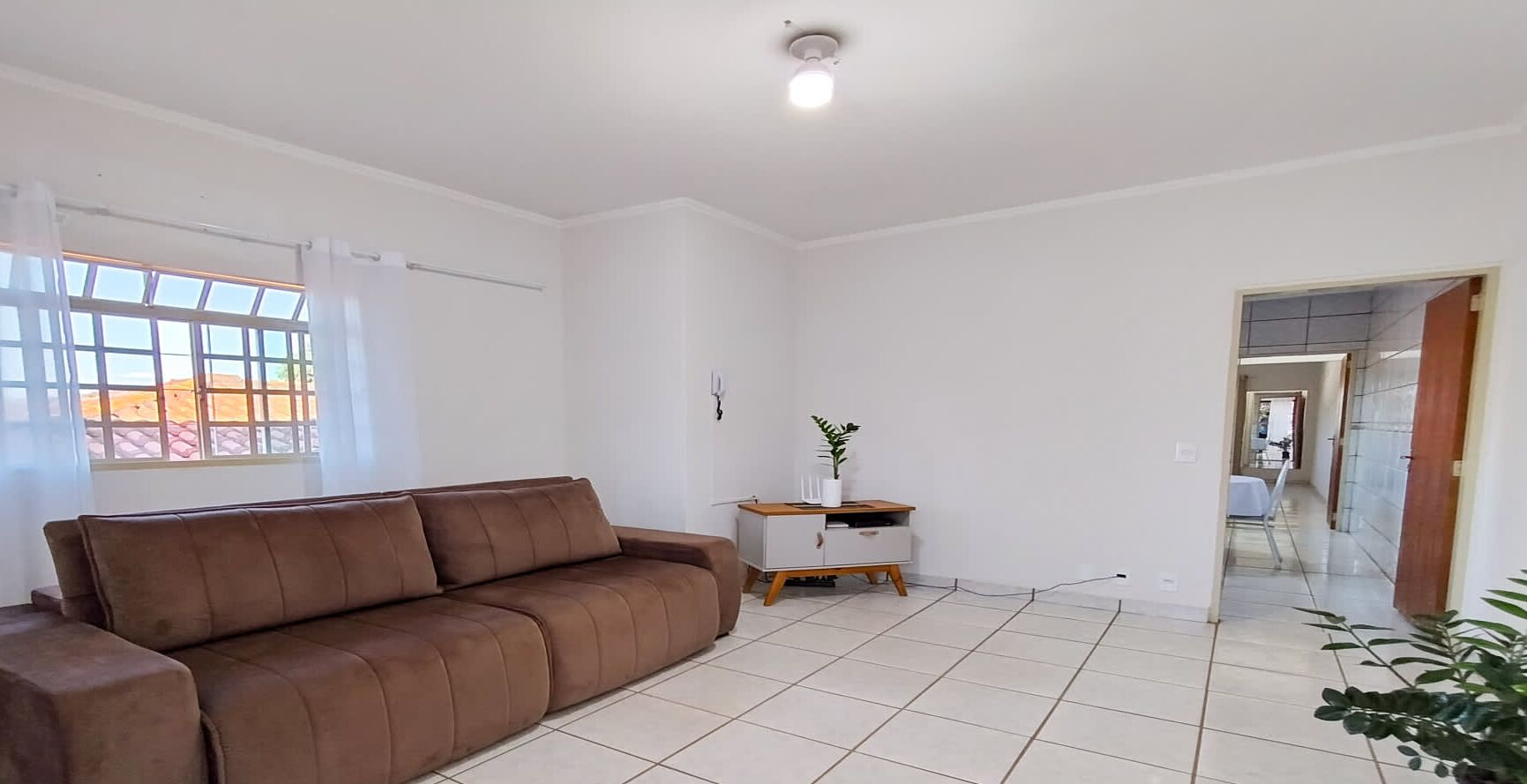 Een ruime woonkamer met een bruine bank, een klein wit bijzettafeltje met een plant, en uitzicht door een raam.
