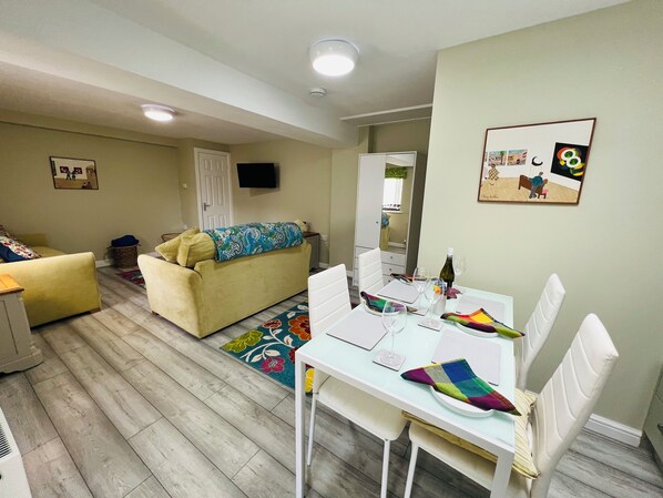 Un salón luminoso con un sofá, una mesa de comedor preparada para dos personas y un espejo que refleja la habitación.