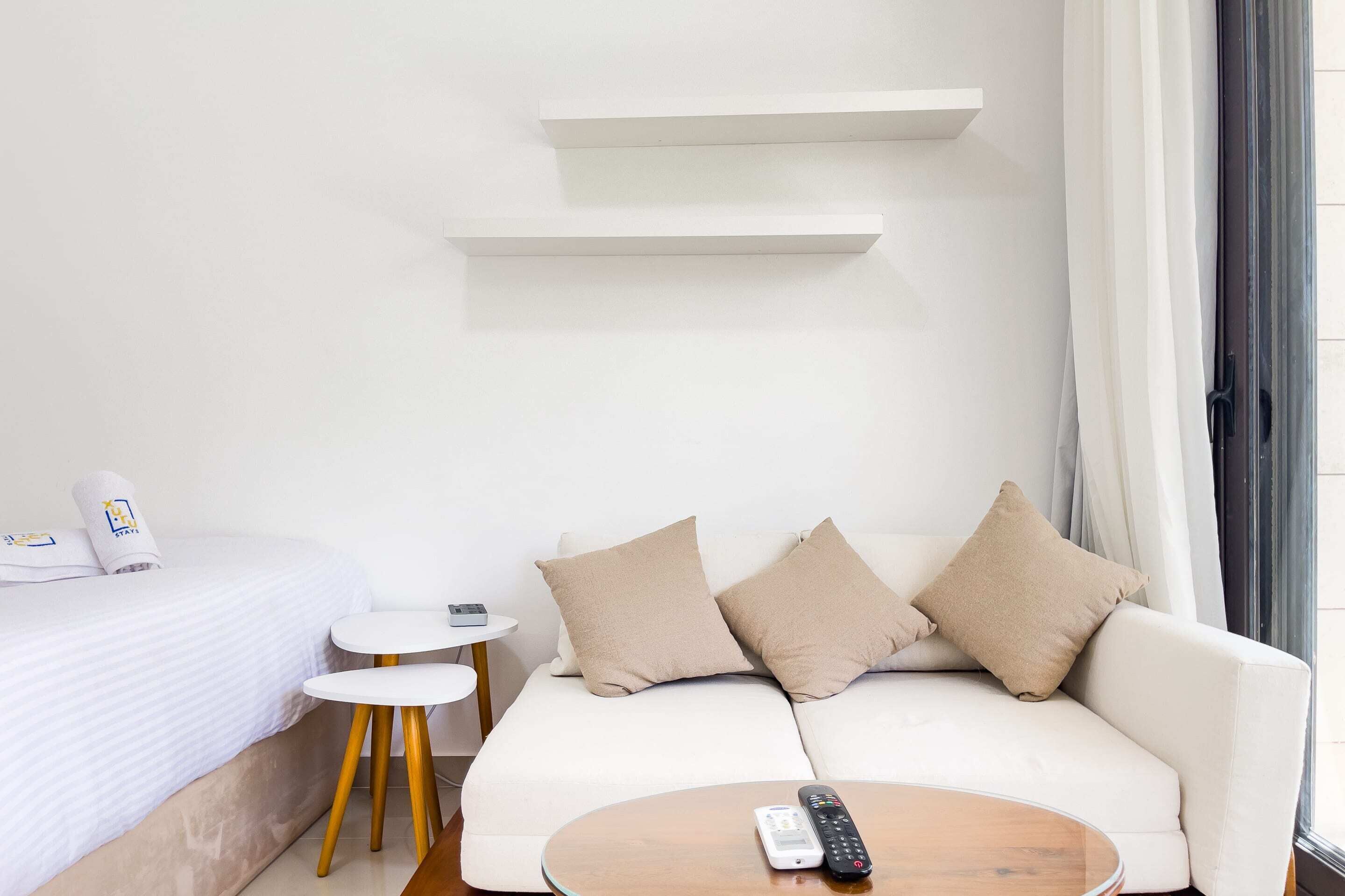 A modern hotel room with a white bed, a white sofa, two beige pillows, a small round wooden table, and a remote control on the table.