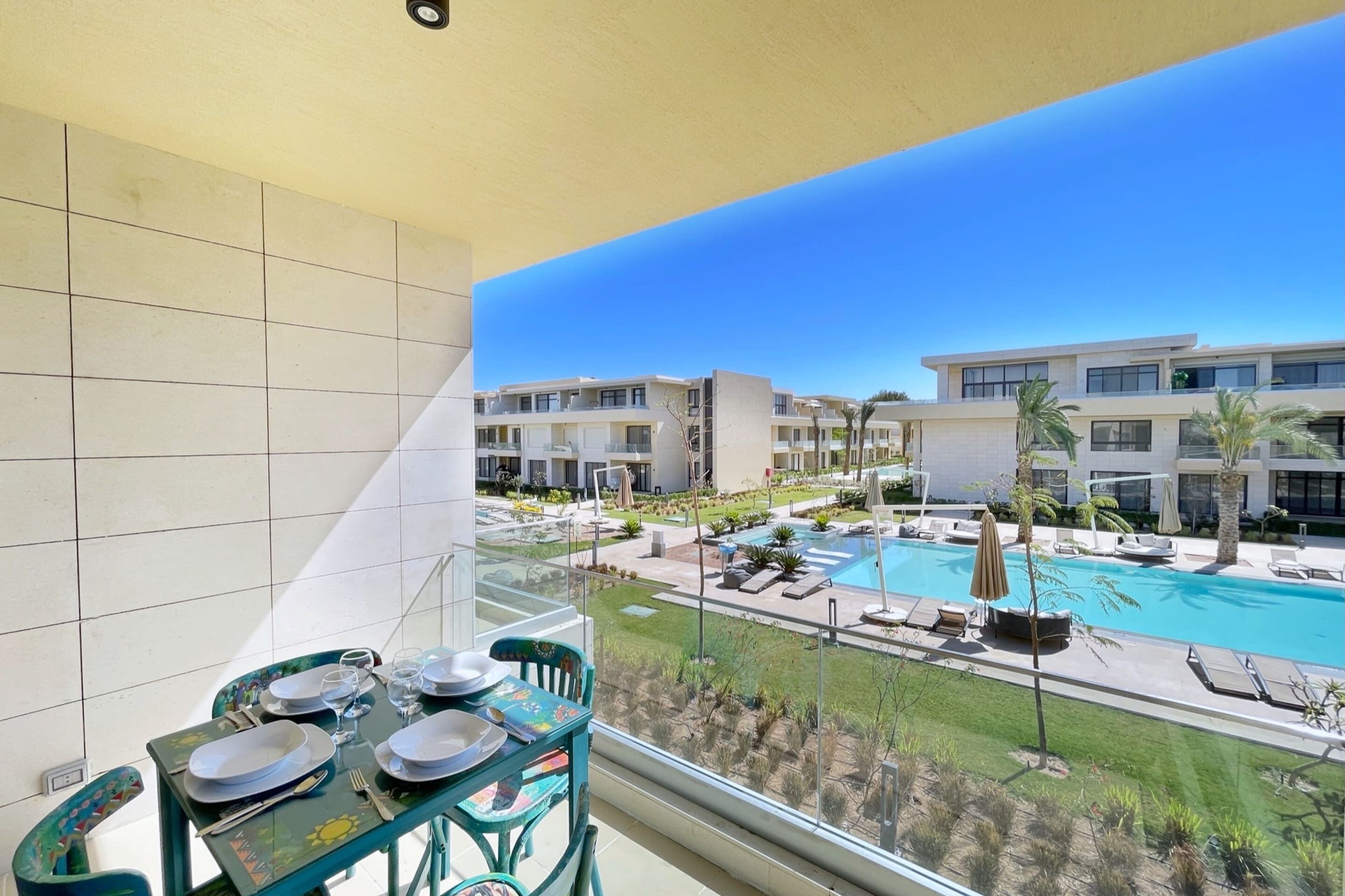 A balcony with a dining table set for four, overlooking a swimming pool and modern buildings.