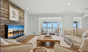 A spacious living room with a flat-screen TV mounted on a stone wall, a wooden coffee table, and a view of the ocean through large sliding doors.