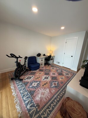 Una habitación con alfombra estampada, una silla azul, una bicicleta estática, un escritorio con lámpara y una puerta blanca.