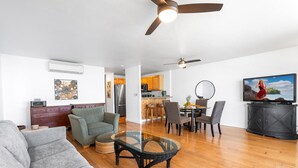 A modern living room with a sofa, armchair, and glass coffee table. There is a dining area with chairs and a TV on a cabinet.