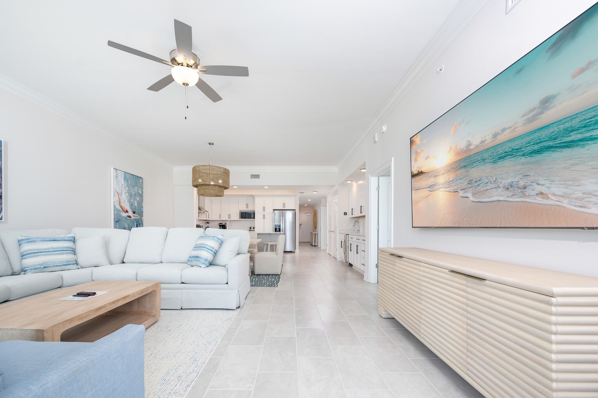 Un salon moderne avec un canapé blanc, une table basse en bois et un grand tableau représentant une scène de plage.