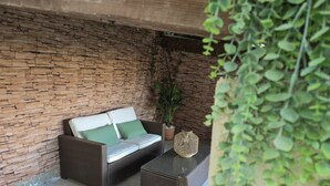 Un canapé en osier avec des coussins verts, une petite table et une plante en pot, le tout installé dans un espace extérieur abrité avec un mur en briques.