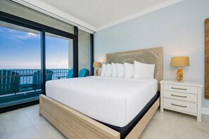 A large bed with a white quilt, a nightstand with a lamp, and a view of the ocean through a balcony. - Phoenix Gulf Shores Ii 1005 4 Bedroom Condo (Gulf Shores)