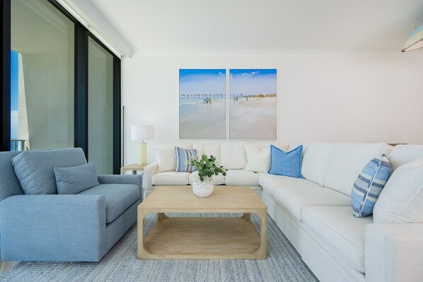 A modern living room with a grey sofa, a white sofa, and a wooden coffee table. There is a large window on the left and two paintings on the wall.