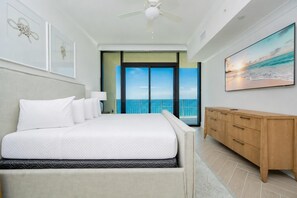 A hotel room with a bed, a wooden dresser, a ceiling fan, and a large window with a view of the ocean.