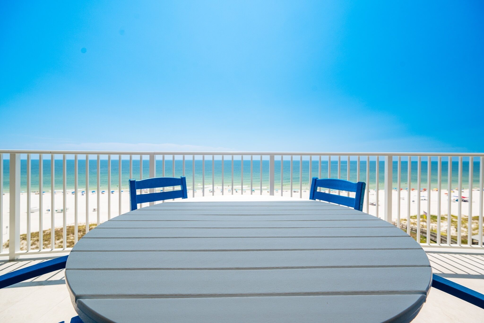 A round table with two blue chairs on a balcony overlooking a beach with white sand and a clear blue sky.