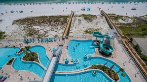 A large outdoor swimming pool with multiple lanes and a water slide, surrounded by beach chairs and umbrellas.