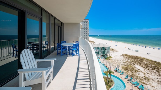 A balcony with white Adirondack chairs, a table, and a pool area with lounge chairs. - Phoenix Gulf Shores Ii 602 3 Bedroom Condo (Gulf Shores)