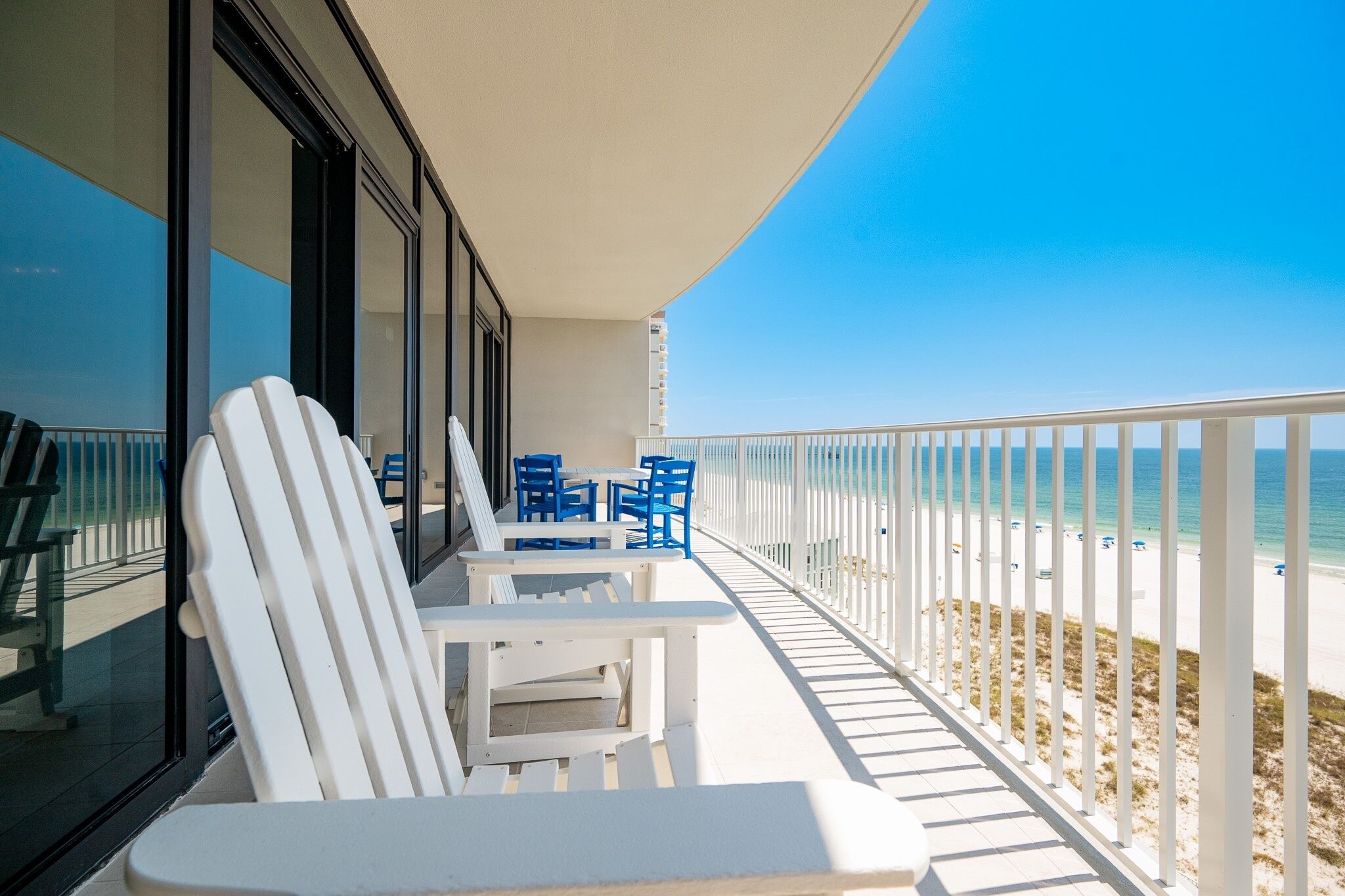 A balcony with white chairs and a table, overlooking a beach with a white railing.