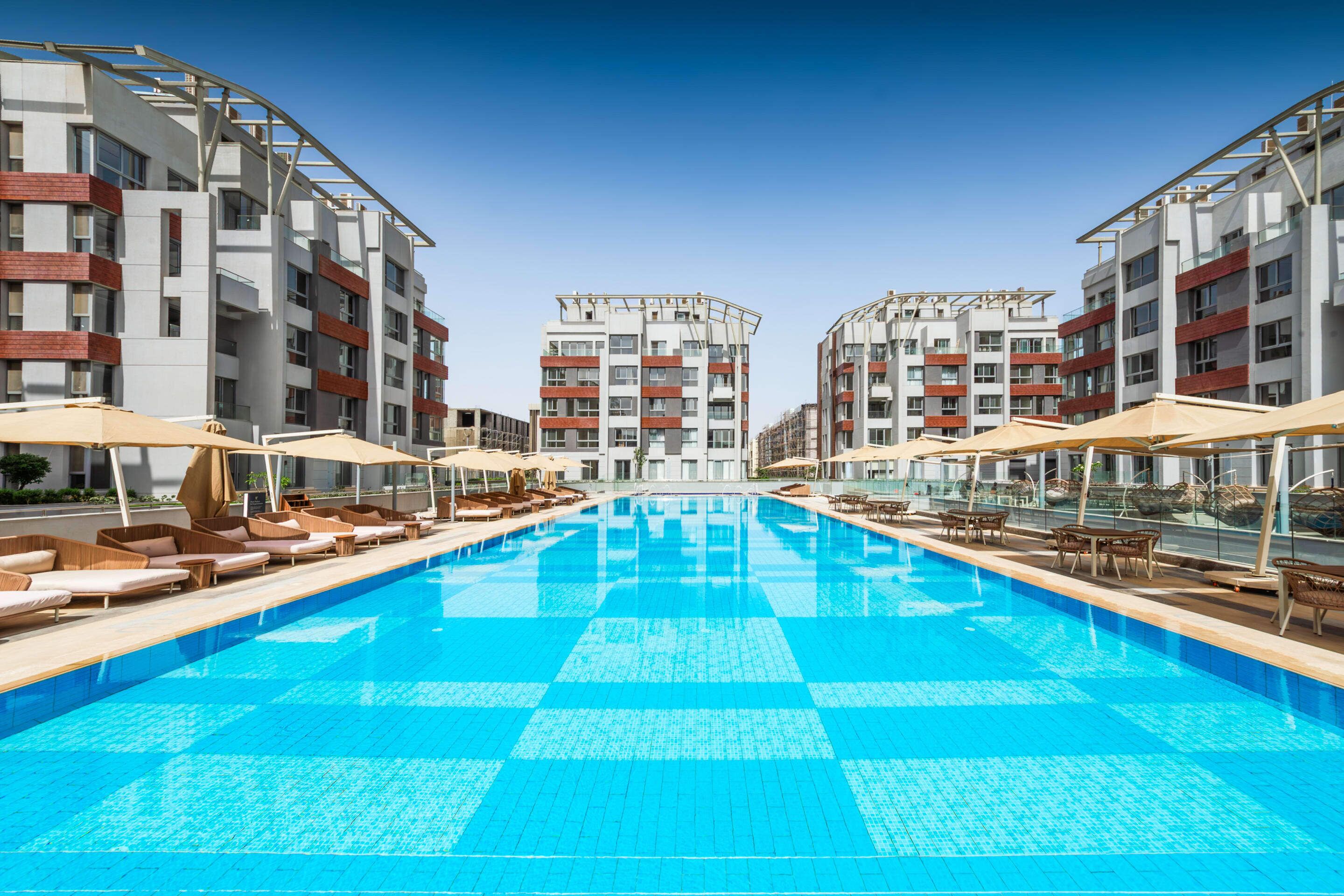 A swimming pool with lounge chairs and umbrellas, surrounded by multi-story buildings with balconies.